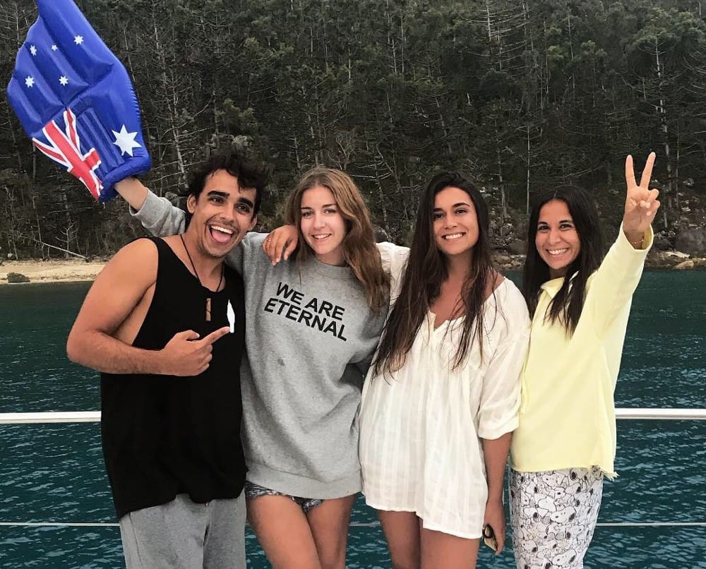 grupo de amigos que sonríen a cámara con una bandera de Australia y están en un barco. Detrás aguas cristalinas
